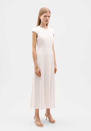 Un vestito bianco aderente, lungo fino al ginocchio, con maniche a cappuccio, tessuto liscio e silhouette dritta. Abbinato a sandali trasparenti con cinturini.