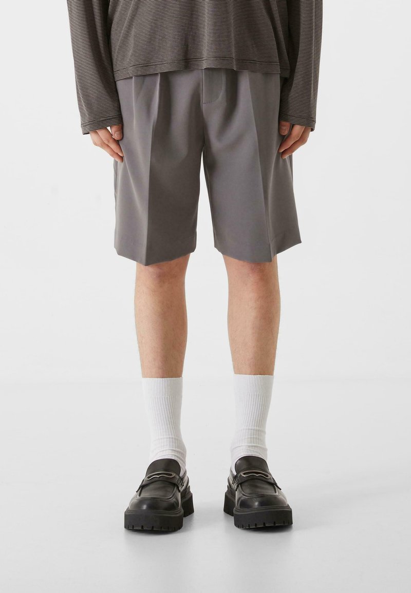 Shorts gris avec des plis, associés à des chaussettes blanches et des chaussures noires. Le haut présente un design à rayures à manches longues dans des tons atténués.