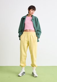 Grøn letvægtsjakke, lyserød cropped top, gule sweatpants med elastisk talje og grå Nike sneakers. Står på en grøn overflade.