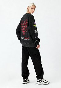 Čierny oversized sveter s červenými srdiečkami, kvetinovými a motýlovými grafikami a textovými detailmi. V kombinácii s čiernymi cargo nohavicami a teniskami.