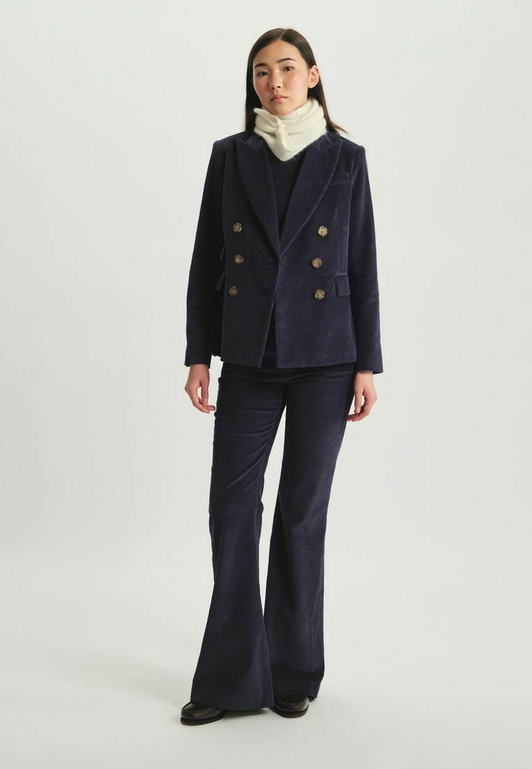 Blazer double boutonné en velours côtelé bleu marine avec des boutons dorés, associé à un pantalon évasé assorti. Une écharpe en crème chaud ajoute un contraste.
