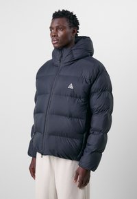 Svart puffjacka med hög krage, quiltad design och dragkedja framtill. Har en liten vit logotyp på bröstet och fickor med dragkedjor.