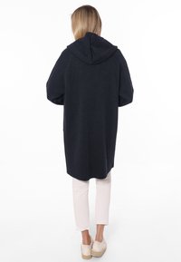 Cappotto lungo navy con cappuccio, maniche oversize, texture a maglia e orlo curvato. Abbinato a pantaloni beige chiaro e espadrillas beige.