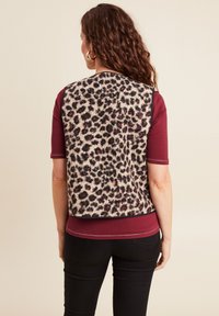 Luipaardprint fleece vest met een afgeronde zoom, zwarte accenten en korte mouwen, gedragen over een bordeauxrode top en gecombineerd met zwarte broeken.