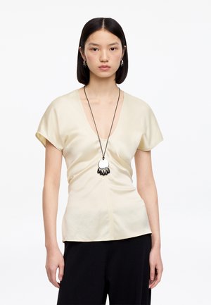 Kobieta o prostych czarnych włosach nosi kremową, krótką bluzkę z dekoltem w serek, czarne spodnie oraz długi naszyjnik z okrągłym wisiorkiem i zwisającymi czarnymi koralikami.