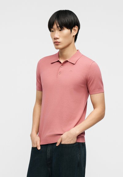 Νέος άνδρας με μαύρα μαλλιά, φορώντας ροζ πόλο μπλουζάκι με κοντά μανίκια και σκούρο παντελόνι, στέκεται με τα χέρια στις τσέπες μπροστά σε απλό φόντο.