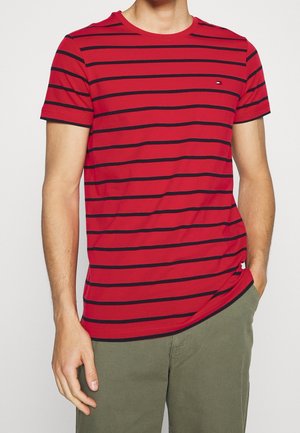 Mężczyzna ubrany w czerwony t-shirt z krótkim rękawem z cienkimi czarnymi poziomymi paskami i zielone spodnie, ręka spoczywa w kieszeni.