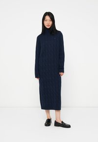 Neulettu tummansininen mekko korkealla kauluksella, letkukuvioinnilla ja pitkillä hihoilla, joka ulottuu polvitaipeeseen. Yhdistetty mustiin loafer-kengiin.