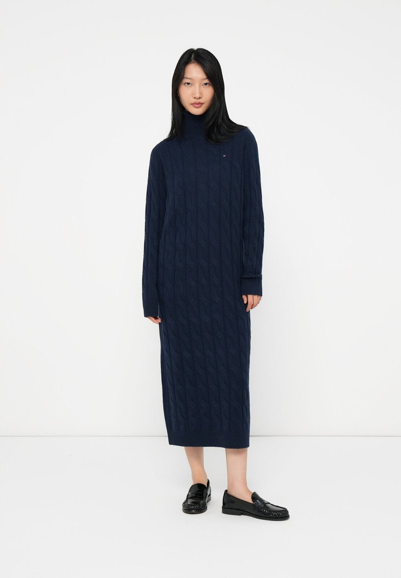 Neulettu tummansininen mekko korkealla kauluksella, letkukuvioinnilla ja pitkillä hihoilla, joka ulottuu polvitaipeeseen. Yhdistetty mustiin loafer-kengiin.