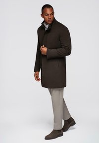 Abrigo de mezcla de lana marrón con un collar alto, bolsillos laterales y una textura suave, combinado con pantalones gris claro y botas marrón oscuro.