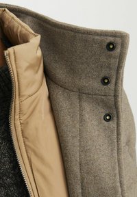 Braune Wollmischung außen mit einem beigen gepolsterten Futter, ausgestattet mit einem hohen Kragen und schwarzen Druckknöpfen, die ein glattes, strukturiertes Finish zeigen.