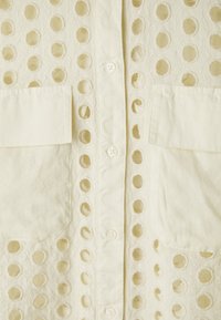 Light yellow blouse with circular cutouts, featuring a button front, two chest pockets, and a textured fabric with embroidered details.