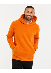 Sudadera de forro polar naranja con bolsillo canguro, capucha grande y logotipo bordado. La tela tiene una superficie suave y texturizada.
