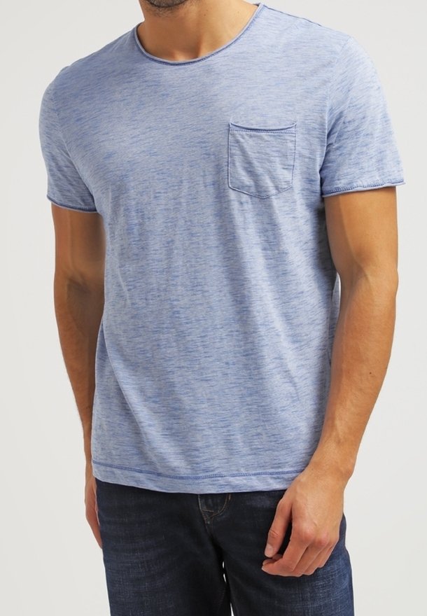 Homme portant un t-shirt bleu clair à manches courtes avec une poche poitrine et un jean bleu foncé, debout devant un fond uni.