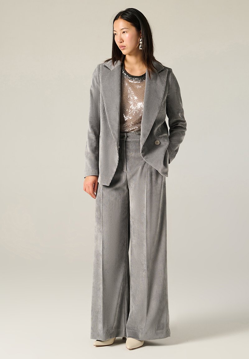 Blazer grigio in corduroy e pantaloni a gamba larga abbinati a un top con paillettes. I dettagli degni di nota includono un colletto, chiusura con bottoni e orecchini a forma triangolare.