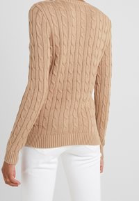 Suéter de cable trenzado en color vainilla con ribete en el dobladillo y los puños, que presenta un patrón texturizado sobre una silueta ajustada. Ideal para el uso casual.