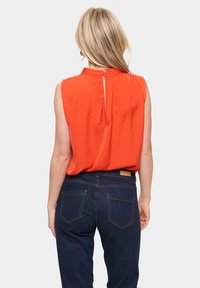 Blusa rossa senza maniche con scollatura arricciata, apertura a chiave sul retro, abbinata a pantaloni in denim blu scuro con due tasche posteriori.