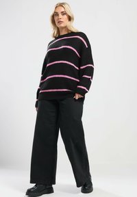 Czarny sweter z dzianiny z różowymi poziomymi paskami, luźny krój i długie rękawy, w połączeniu z szerokimi czarnymi spodniami i botkami sięgającymi kostki.