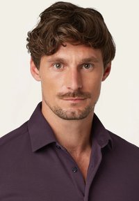 Lila Polohemd mit Kragen, Knopfleist und strukturiertem Stoff, getragen von einem Mann mit kurzen, welligen Haaren und Gesichtshaar.