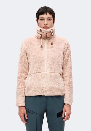 Personne portant une veste rose pâle duveteuse à fermeture éclair avec col montant et cordons, associée à un pantalon bleu foncé avec poches.