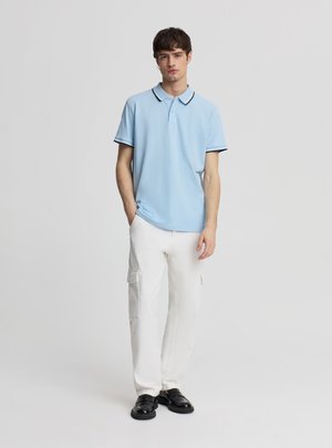 Giovane uomo in piedi con le mani parzialmente nelle tasche, indossa una polo celeste, pantaloni cargo bianchi e scarpe nere su sfondo neutro.