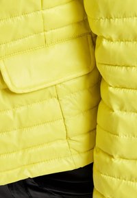 Yellow quilted jacket with a smooth texture, featuring a flap pocket and horizontal stitching across the fabric.