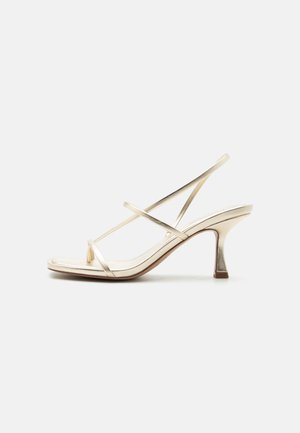 Sandale métallique beige à bout ouvert avec fines brides croisées et talon mi-haut effilé sur un fond clair uni.