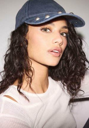 Młoda osoba o kręconych ciemnych włosach, nosząca niebieską czapkę i białą prążkowaną koszulkę, patrząca lekko w górę na jednolitym tle.