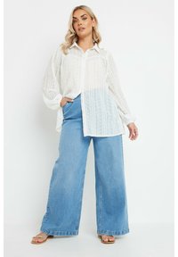 Camicia bianca a maniche lunghe con bottoni e motivo tessuto, abbinata a jeans a vita alta a gamba larga di colore blu chiaro e sandali flat.