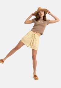 Cămașă maro cu mânecă scurtă și grafică albă, combinată cu pantaloni scurți raiți galbeni. Modelul poartă o pălărie bej și sandale cu talpă groasă maro.