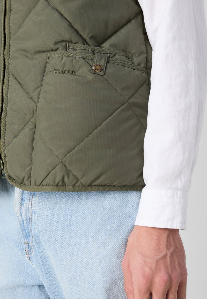 Olivgrüne gesteppte Weste mit Fronttasche, Barbour-Logo-Detail und strukturiertem Finish, getragen über einem weißen Langarmshirt.