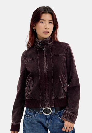 Jeune femme aux cheveux rouge foncé portant une veste en velours côtelé violet foncé, un jean bleu et des bagues argentées aux doigts sur un fond uni.