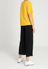 Keltainen puuvillaitaidetta lyhythihaisissa t-paidassa yhdistettynä hajavalta mustiin leveälahkeisiin housuihin ja valkoisiin urheilukenkiin. Rentoa vaatetusta, neutraali tausta.