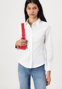 Cămașă albă cu guler și mâneci lungi, asortată cu blugi albaștri. Ținând o geantă roșie din piele cu un design structurat și curea.