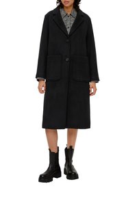 Schwarzer Wollmischmantel mit Reverskragen, zwei vorderen Taschen und einem einzigen Knopfverschluss, kombiniert mit schwarzen Stiefeletten.