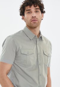 Camisa de botones de manga corta en gris claro con dos bolsillos en el pecho, cuello puntiagudo y una textura de tela suave.