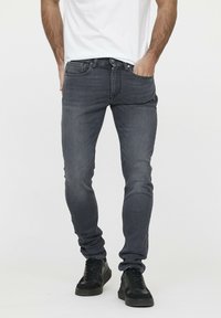 Schmale, dunkelgraue Jeans mit glatter Textur, fünf Taschen und dezentem Bleichen. Getragen mit einem weißen T-Shirt und schwarzen Turnschuhen.