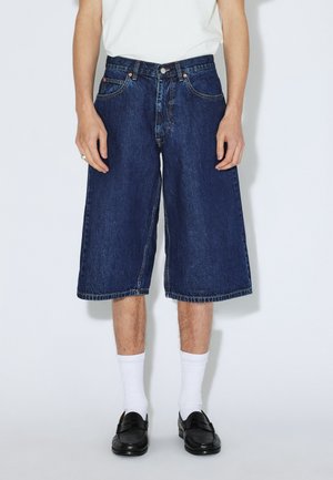 Shorts en jean, bleu foncé, longueur genou avec une coupe ample. Comprend cinq poches, des coutures visibles et une fermeture par bouton à la taille.