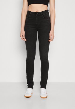 Personne portant un jean skinny noir taille haute, des baskets blanches et un crop top noir, debout devant un fond gris clair uni.