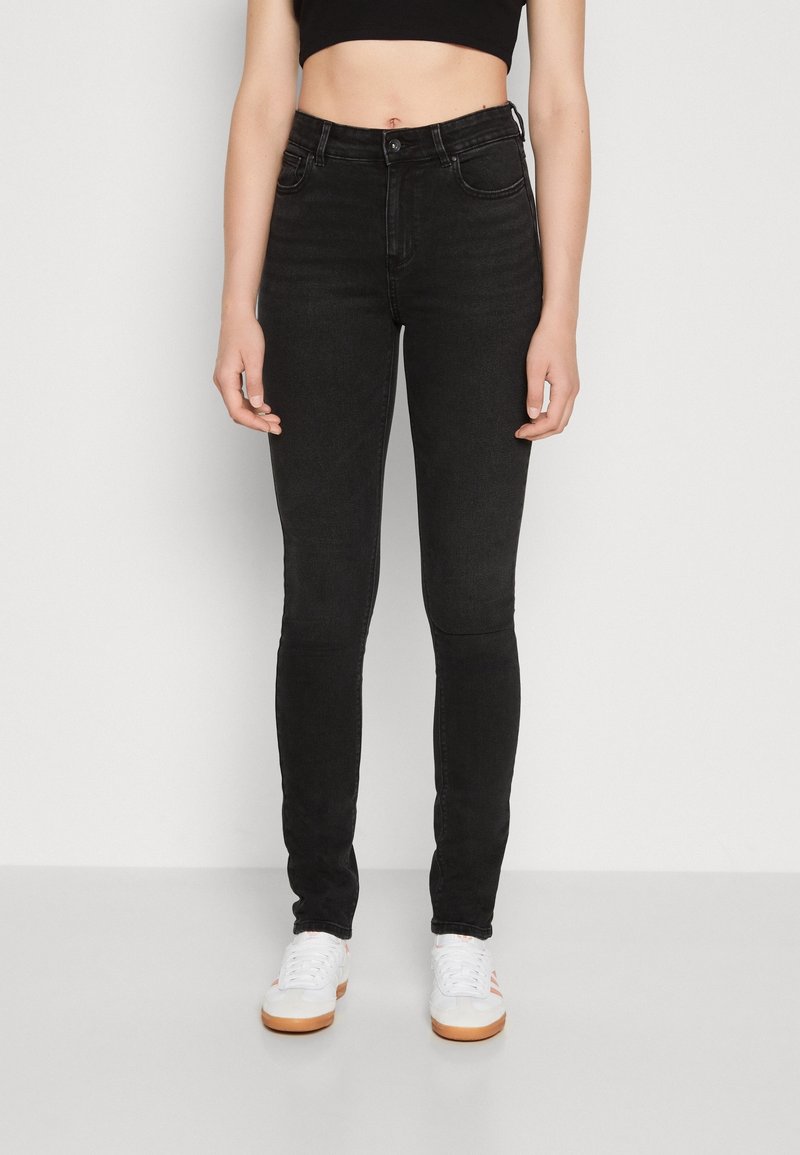 Personne portant un jean skinny noir taille haute, des baskets blanches et un crop top noir, debout devant un fond gris clair uni.