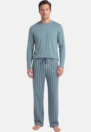 Man staat op blote voeten, draagt een lichtblauw hemd met lange mouwen en lichtblauwe pyjamabroek met marineblauwe verticale strepen.