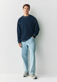 Granatowa koszula z długim rękawem z kontrastowymi jasnymi szwami, zestawiona z jasnoniebieskimi prostymi spodniami dżinsowymi i szarymi sneakersami. Minimalistyczny design.