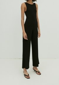 Mono negro de canalé con diseño sin mangas, piernas anchas y cintura ajustada. Combinado con chanclas negras, que muestran una textura suave.