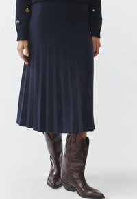 Marineblauwe plissé midi-rok met een gestructureerde weving, gecombineerd met bruine leren cowboylaarzen met spitse neuzen en schuine hakken.