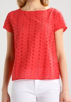 Woman wearing a coral short-sleeve blouse with floral eyelet pattern paired with white pants against a plain background.