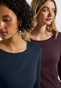Dvě ženy na sobě mají dlouhé rukávy, jedna v námořnické modři a druhá ve tmavě burgundské. Topy mají kulaté výstřihy a přiléhavé střihy.