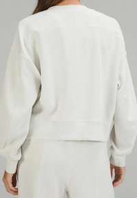 Persona vistiendo un sudadera de manga larga color blanco roto y pantalones a juego, mostrada de espaldas contra un fondo gris liso.