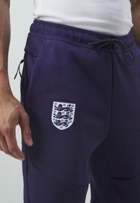 Lila joggers med midjeband och dragsko, sidofickor och en broderad Englands-sköld med lejon och rosor. Mjuk textur, sportig design.