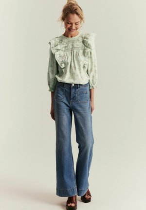 Kvinde smiler nedad iført en lys grøn blomstret bluse med flæser, højtaljede vidde-ben jeans og brune åbne platformssko.