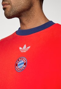 Κόκκινη αθλητική φανέλα με σκούρο μπλε ριμπ λαιμόκοψη, με λευκό λογότυπο Adidas και κεντημένο σήμα του FC Bayern München.